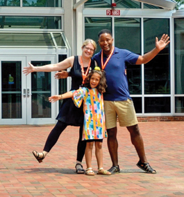 A mother, father and young daughter stand together with their arms outstretched in front of the Brandeis library